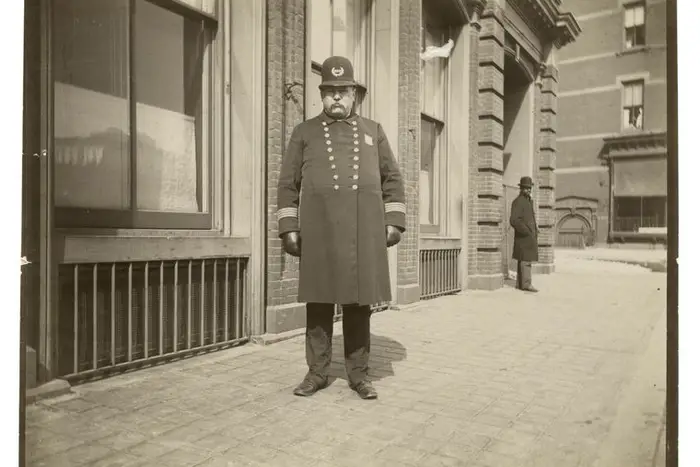 A sepia photo of a NYC police officer with a belly and wearing a rounded hat